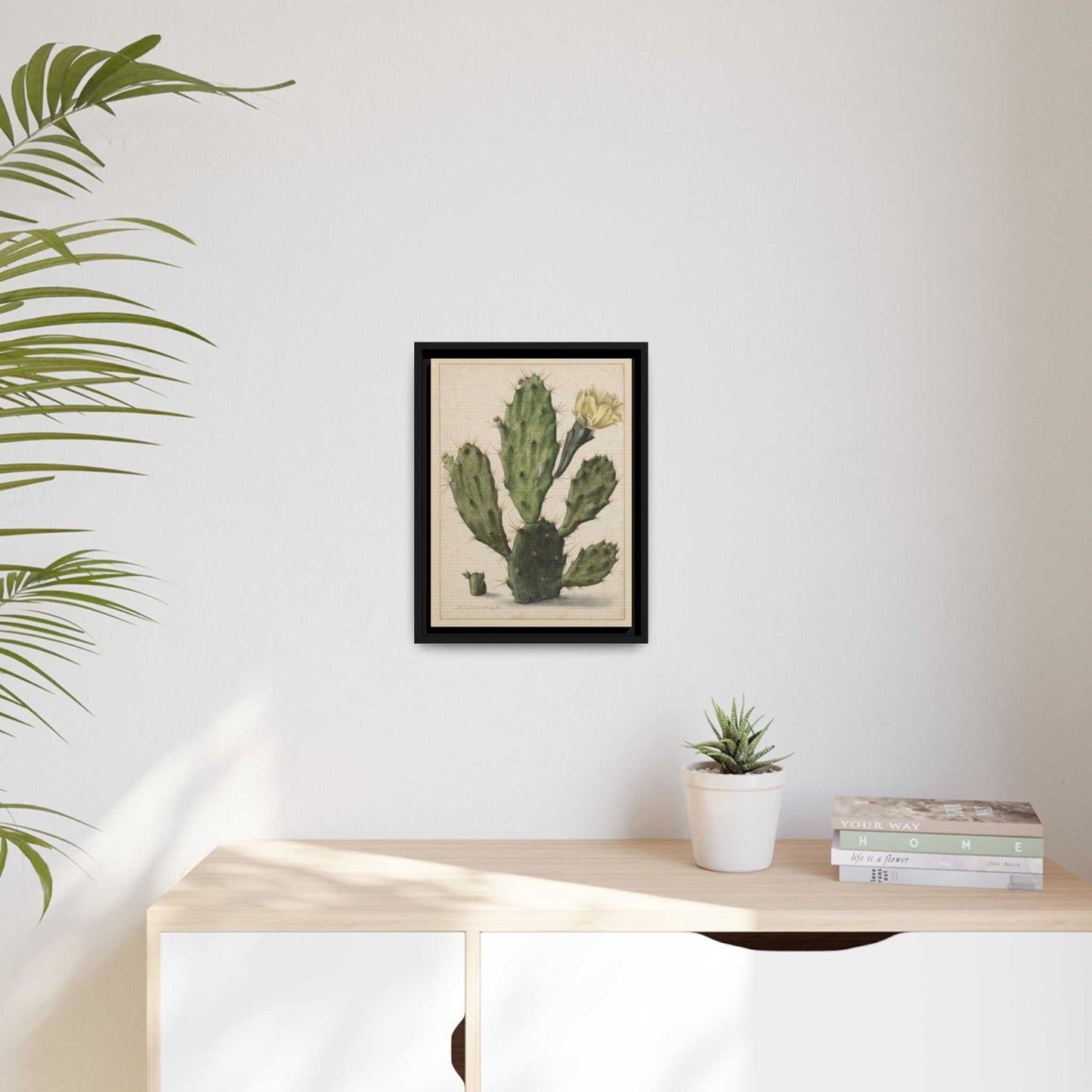 Cactus with yellow flower - black framed poster. Pictured on a wall above a white credenza. A plant sits to the left side
