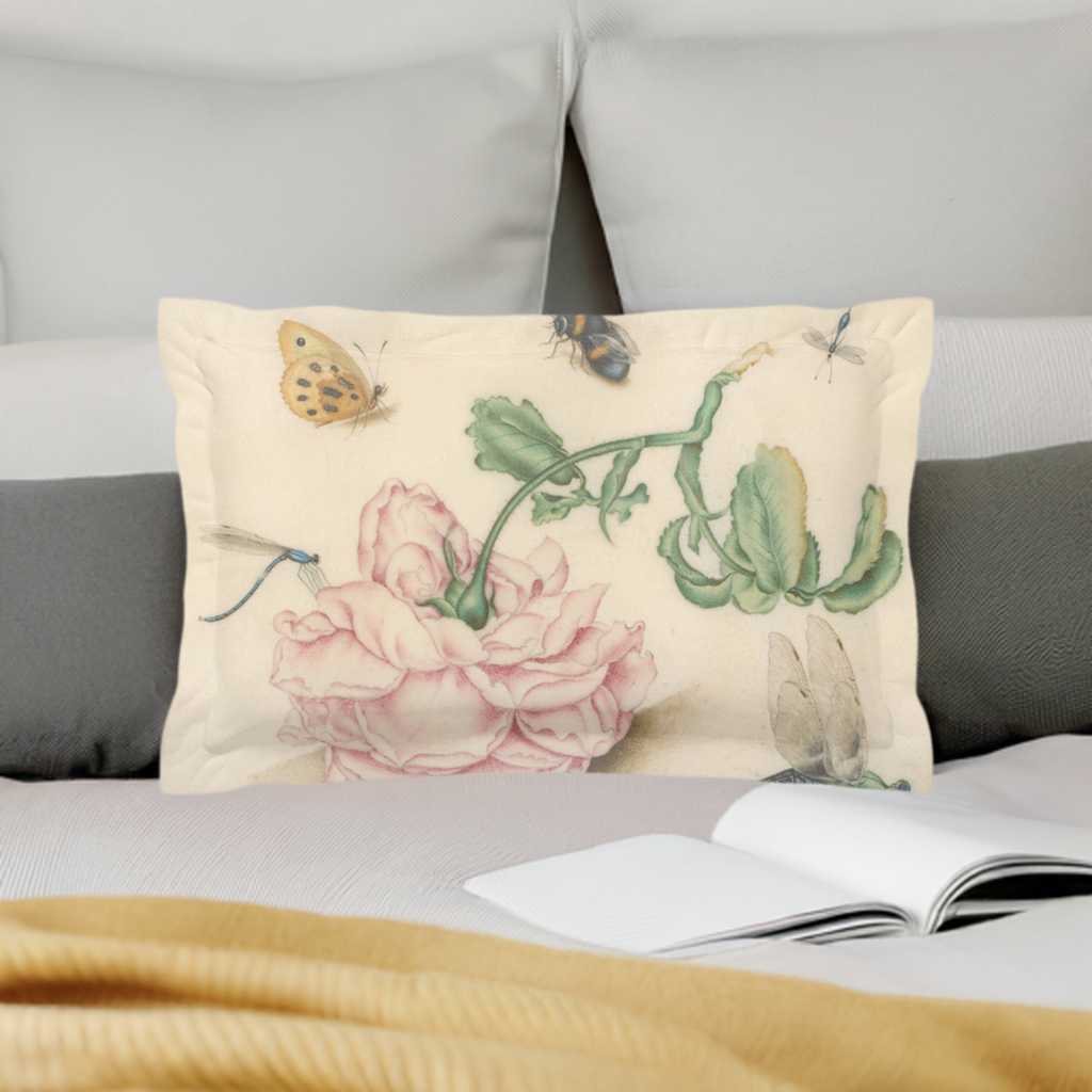 A micro fibre Pillow sham with 2 inch flange. A botanical print of butterflies , a bee and blue dragonflies. A large pink downturned rose is at the centre of the print. Pictured in front of a grey head board on a white duvet cover with a golden cable knit blanket and an open book on the bed