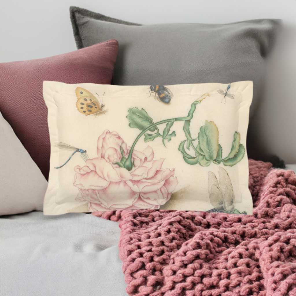 A micro fibre Pillow sham with 2 inch flange. A botanical print of butterflies , a bee and blue dragonflies. A large pink downturned rose is at the centre of the print. Pictured on white bedding and a pink cable knit blanket with pink and green pillows behind it.