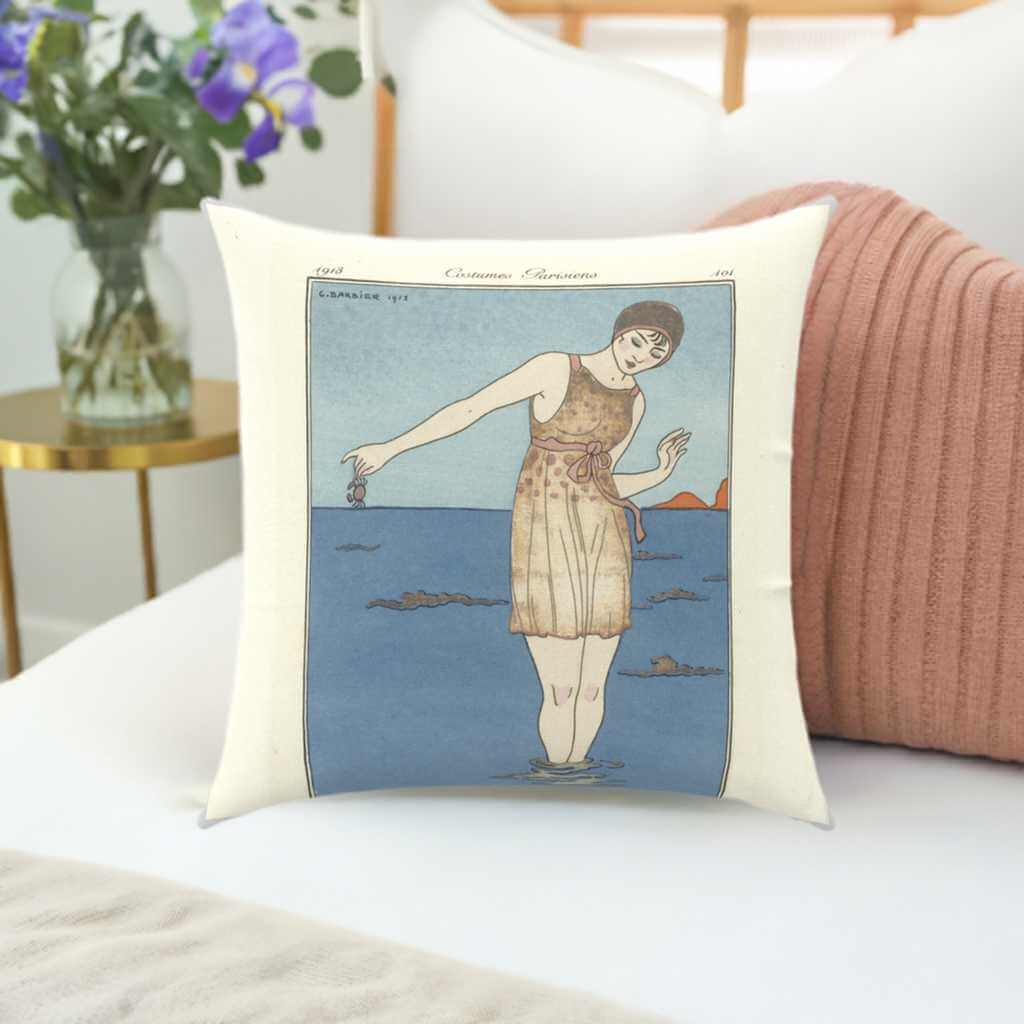 Beach Babe 1913' Throw Pillow 100% Cotton Twill in a 16x16 inch double-sided print cushion with poly fill insert and Zipper for easy removal and washing. Vintage illustration of woman in an antique bathing suit. On a blue background with a cream border. Pictured on a white duvet cover in front of a knit salmon coloured cushion. a brass bedside table is beside the bed with a vase of flowers