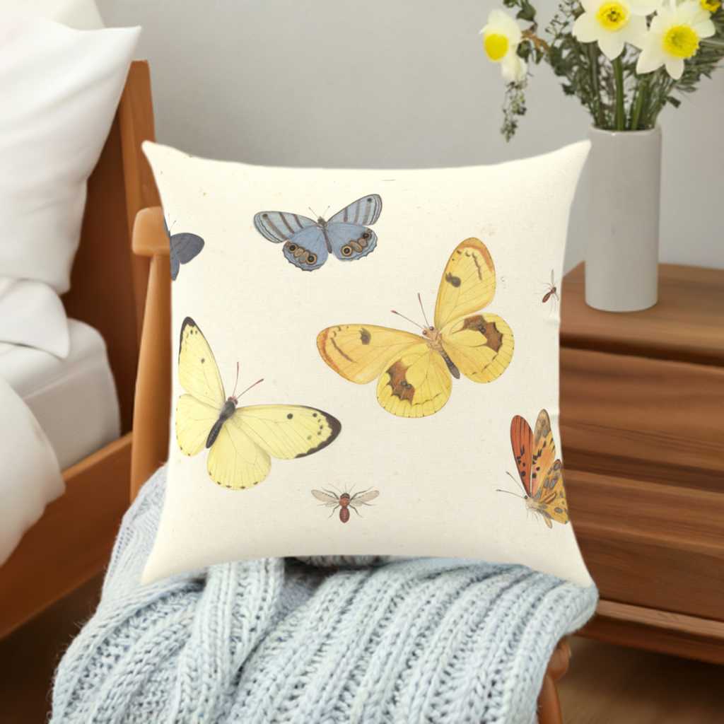 a twill cotton throw pillow with botanical style depictions of blue, yellow and orange butterflies on a cream background on a bed with a white duvet cover. a wooden side table to the left holds a vase of eucalyptus leaves