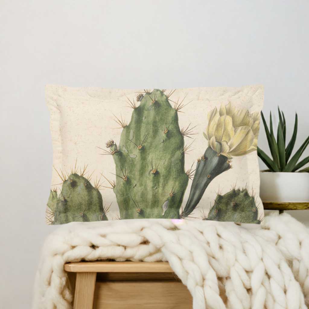 Pillow sham with 2 inch flange - micro fibre - Cactus with yellow flower on cream background - Pictured in front of a green velvet headboard on a white duvet cover behind a yellow knit blanket lying on top of a wooden night stand. A plant sits to the right of the pillow sham on the night stand