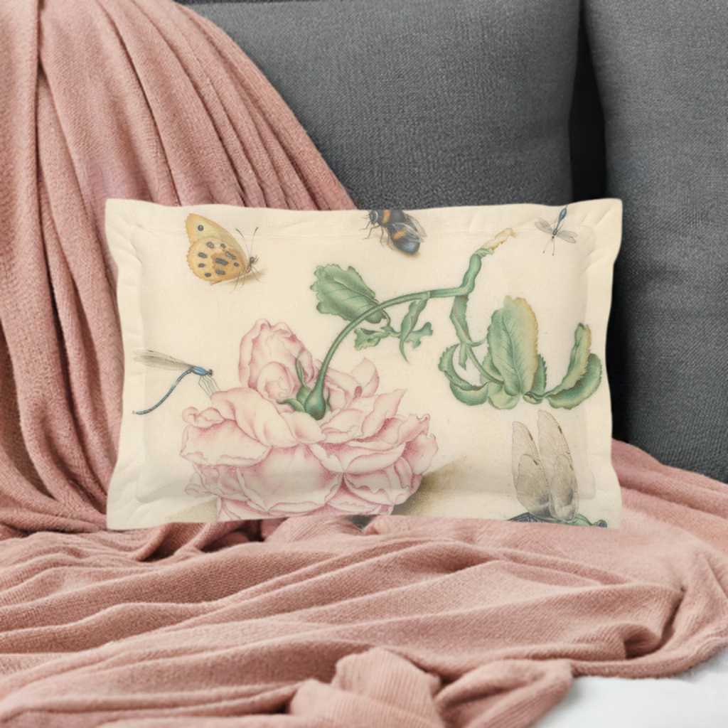 A micro fibre Pillow sham with 2 inch flange. A botanical print of butterflies , a bee and blue dragonflies. A large pink downturned rose is at the centre of the print. It is pictured on a pale pibk cable knit blanket that is draped over a gray chair