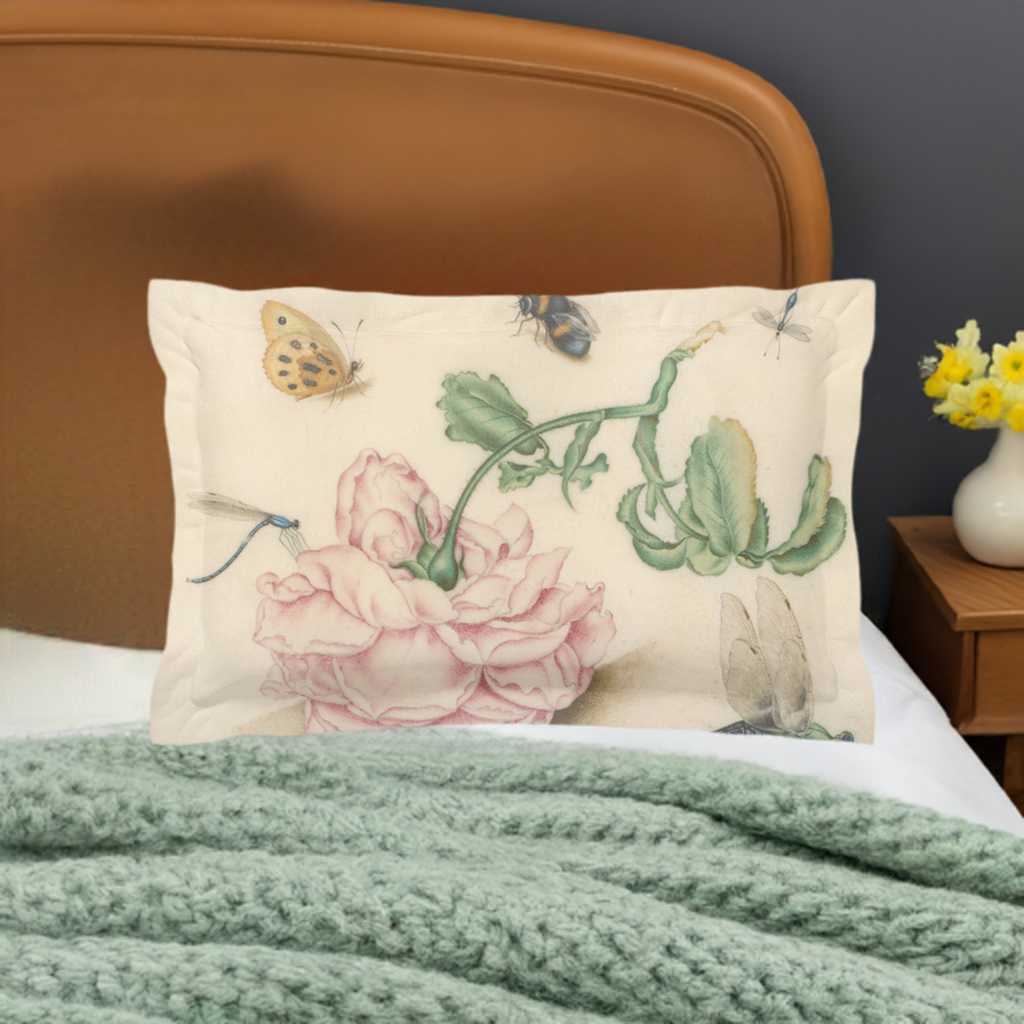 A micro fibre Pillow sham with 2 inch flange. A botanical print of butterflies , a bee and blue dragonflies. A large pink downturned rose is at the centre of the print. Pictured in front of a honey coloured wooden headboard and a pale green cable knit blanket in front. A vase of flowers sits on the side table